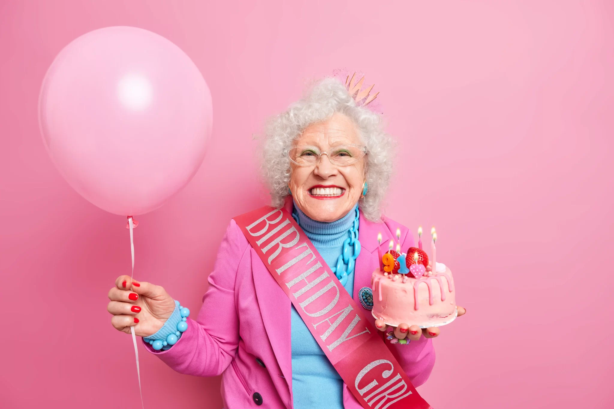 studio-shot-of-happy-wrinkled-female-pensioner-with-bright-makeup-smiles-toothily-holds-festive-cake-with-burning-candles-has-festive-mood-carries-inflated-balloon